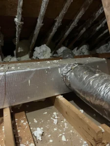 Foil-wrapped ventilation ducts running through a loft space with exposed wooden rafters and insulation debris in Cheshire