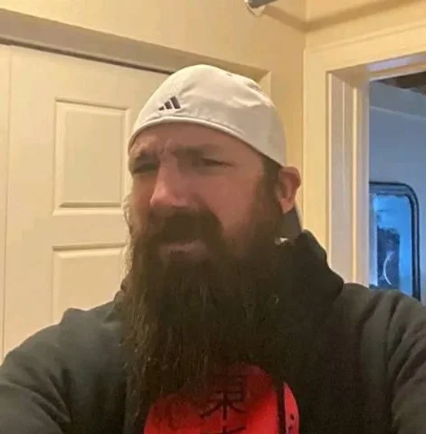 A portrait of a smiling middle-aged man with short brown hair and glasses, wearing a tan Carhartt pocket t-shirt. He is sitting outdoors in front of a glass partition with a green field and trees in the background, used for a property service review in Banbury.