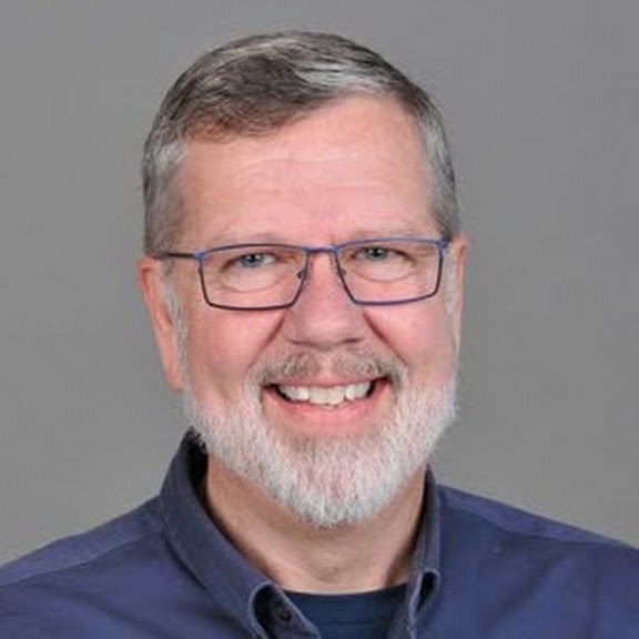 An outdoor selfie of a smiling male customer with dark messy hair and earbuds, wearing a grey unbuttoned shirt and a black bag strap, standing in a car in Basildon, Essex, after a successful loft insulation removal project.