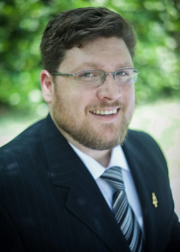 A studio-style portrait of a professional-looking man with short grey hair and glasses, wearing a light blue checkered shirt against a pale blue background.