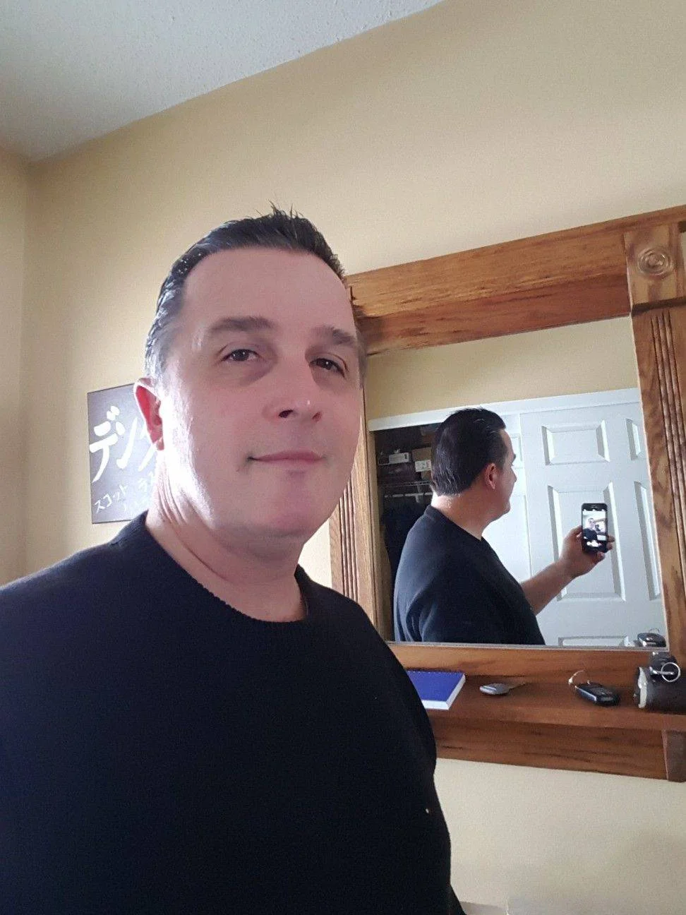 A man in a black sweater taking a mirror selfie with his smartphone. The wooden mirror frame and Japanese wall art are visible. Used as a residential review for a period property owner in Chester needing professional loft insulation extraction.