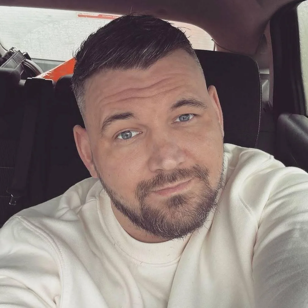 A close-up selfie portrait of a middle-aged man with short dark hair, blue eyes, and a beard, wearing a cream-white sweatshirt. He is sitting inside a car looking directly at the camera with a direct expression, used as a customer testimonial in Cirencester.