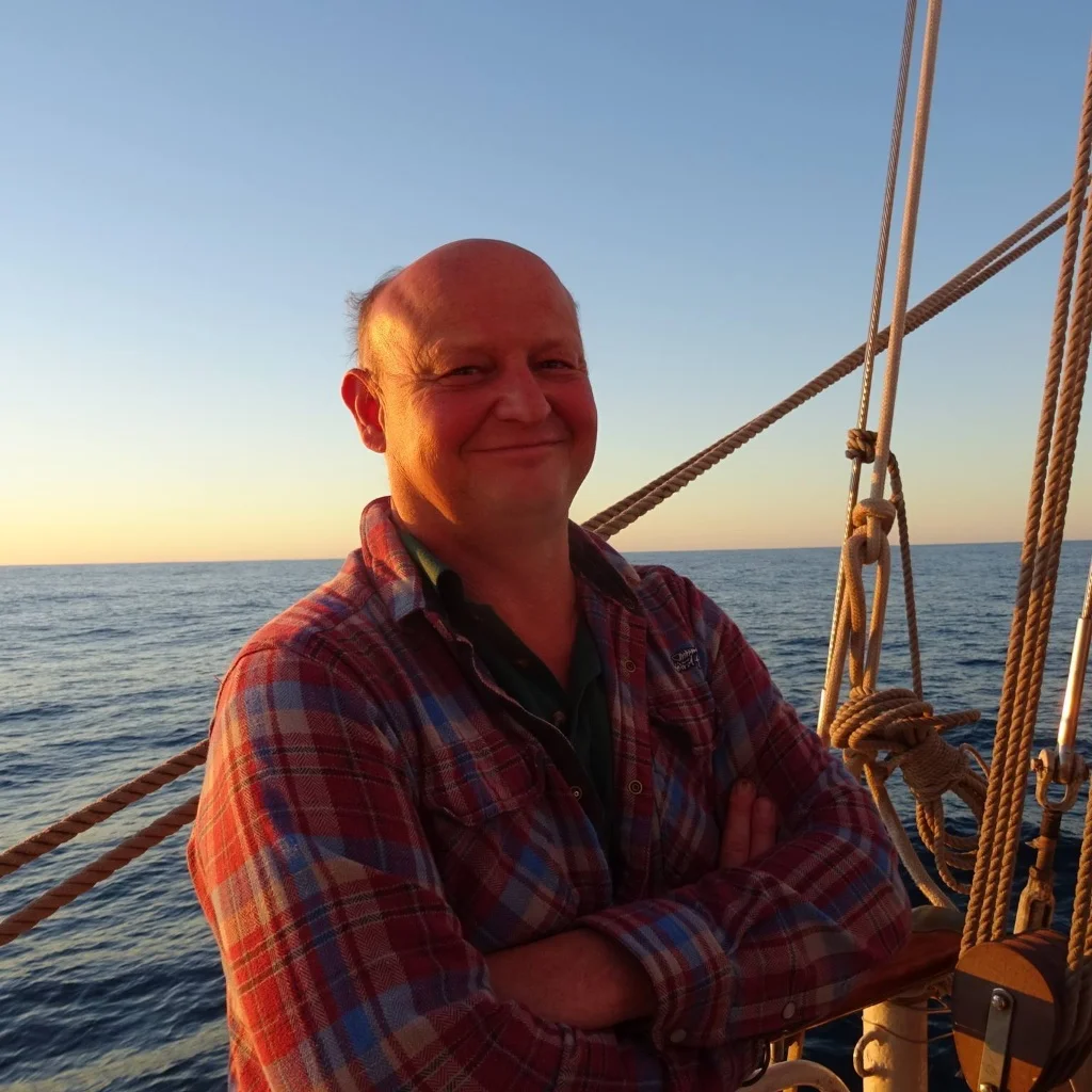 Outdoor portrait of a smiling bald senior man with crossed arms, wearing a red and blue plaid flannel shirt, standing on a sailing boat at sunset with the sea in the background, providing a review from Ely.