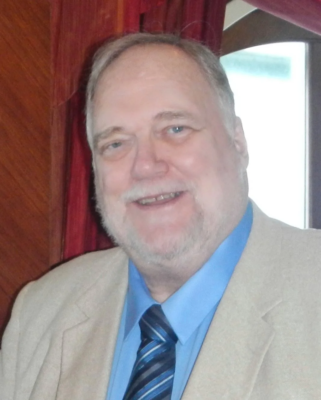 A portrait of a smiling senior man with grey hair and a goatee, wearing a tan suit jacket and a blue shirt with a striped tie. Used as a professional customer testimonial for spray foam extraction in a Victorian villa in Torquay.
