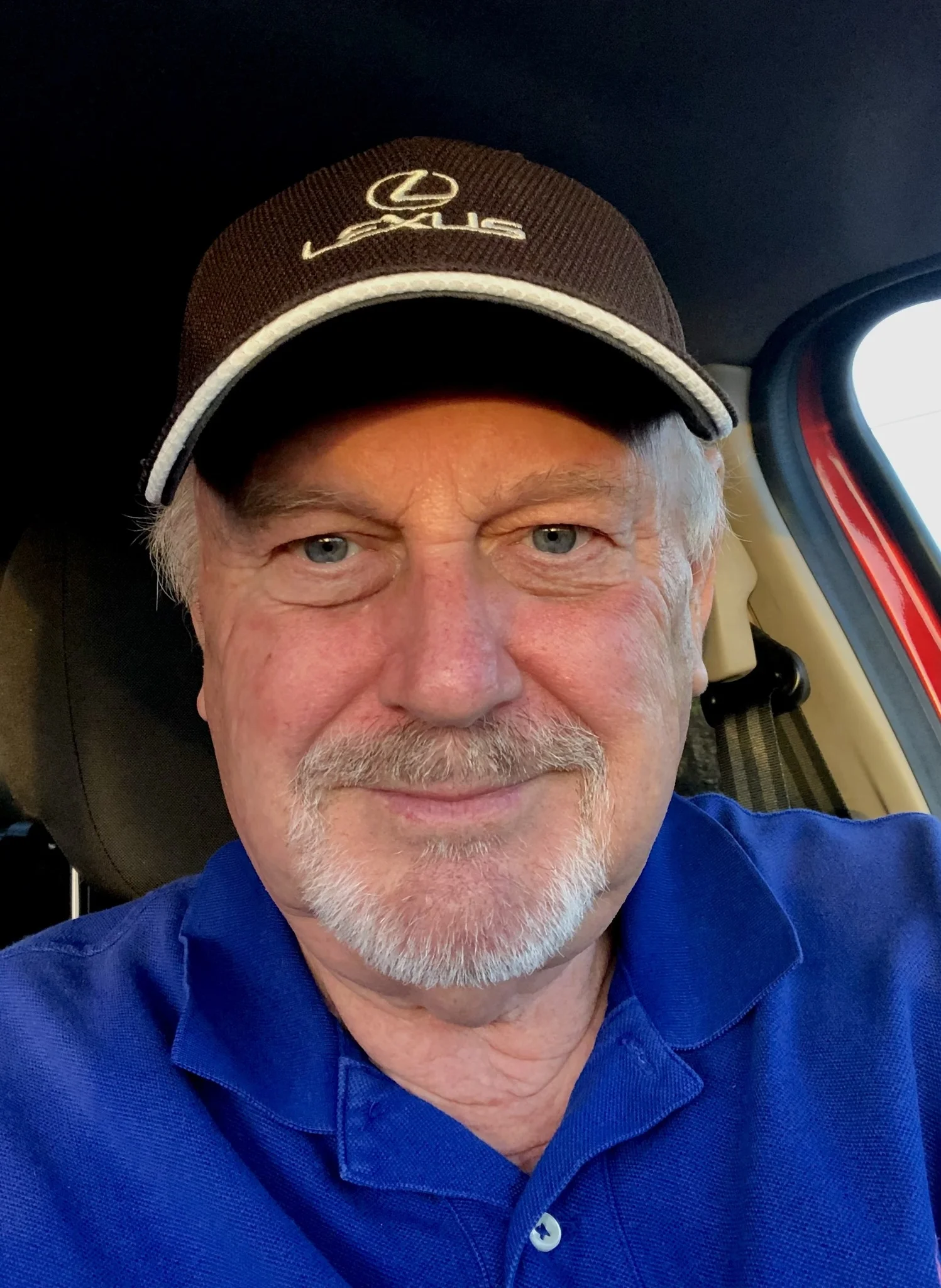 A smiling senior man with a grey beard and blue eyes, wearing a brown Lexus baseball cap and a blue polo shirt, sitting in a car. This image represents a satisfied resident in Exmouth who had spray foam removed for mortgage compliance.