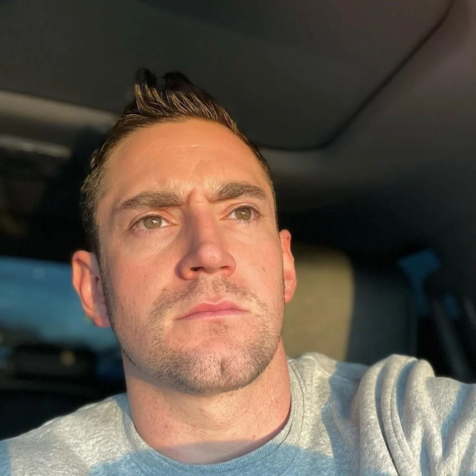 A close-up selfie portrait of a serious young man with short brown spiky hair, wearing a light grey crewneck t-shirt. Strong, direct sunlight illuminates his face. He is looking up and slightly to the side with a serious expression, used as a customer testimonial in Gloucester.
