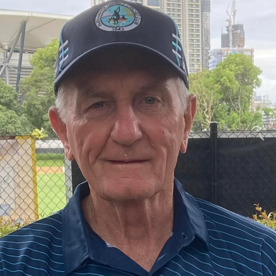 A portrait of a senior man wearing a navy blue 'Parramatta District Cricket Club' cap and a striped blue polo shirt. He is standing outdoors in front of a chain-link fence with trees and city buildings in the background, used for a property service review in Grantham.