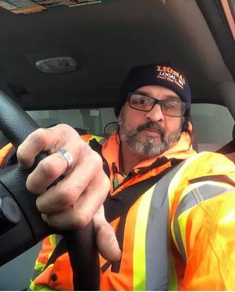 A middle-aged man with a beard, wearing prescription glasses and a high-visibility orange/yellow reflective jacket over a dark top. He is actively steering a vehicle with his left hand, and a large silver ring is visible on his index finger. This is a local Huyton trade expert, used to authorize a residential review for spray foam removal in Prescot, Merseyside.