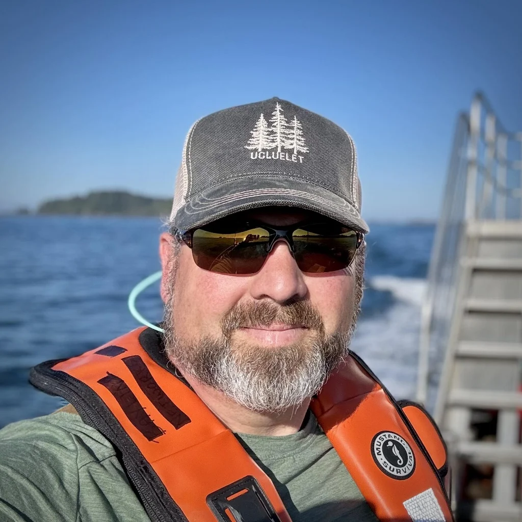 Outdoor portrait of a smiling middle-aged man with short hair and a white beard, wearing a grey ‘Ucluelet’ trucker hat, green t-shirt, sunglasses, and an orange life vest, standing on a boat with the blue sea and distant islands. Used as a customer testimonial for insulation services in King's Lynn, Norfolk.