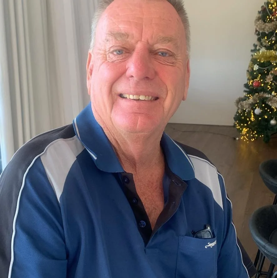 A close-up portrait of a smiling senior man with short grey hair and blue eyes, wearing a blue and grey patterned polo shirt. He is sitting indoors with a festive tree in the background, used as a customer testimonial for insulation services in Lincoln.