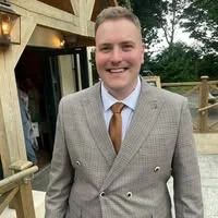 A smiling young man in a tan checked double-breasted suit and gold tie, standing outside a wooden building, providing a testimonial for property services in Loughborough.