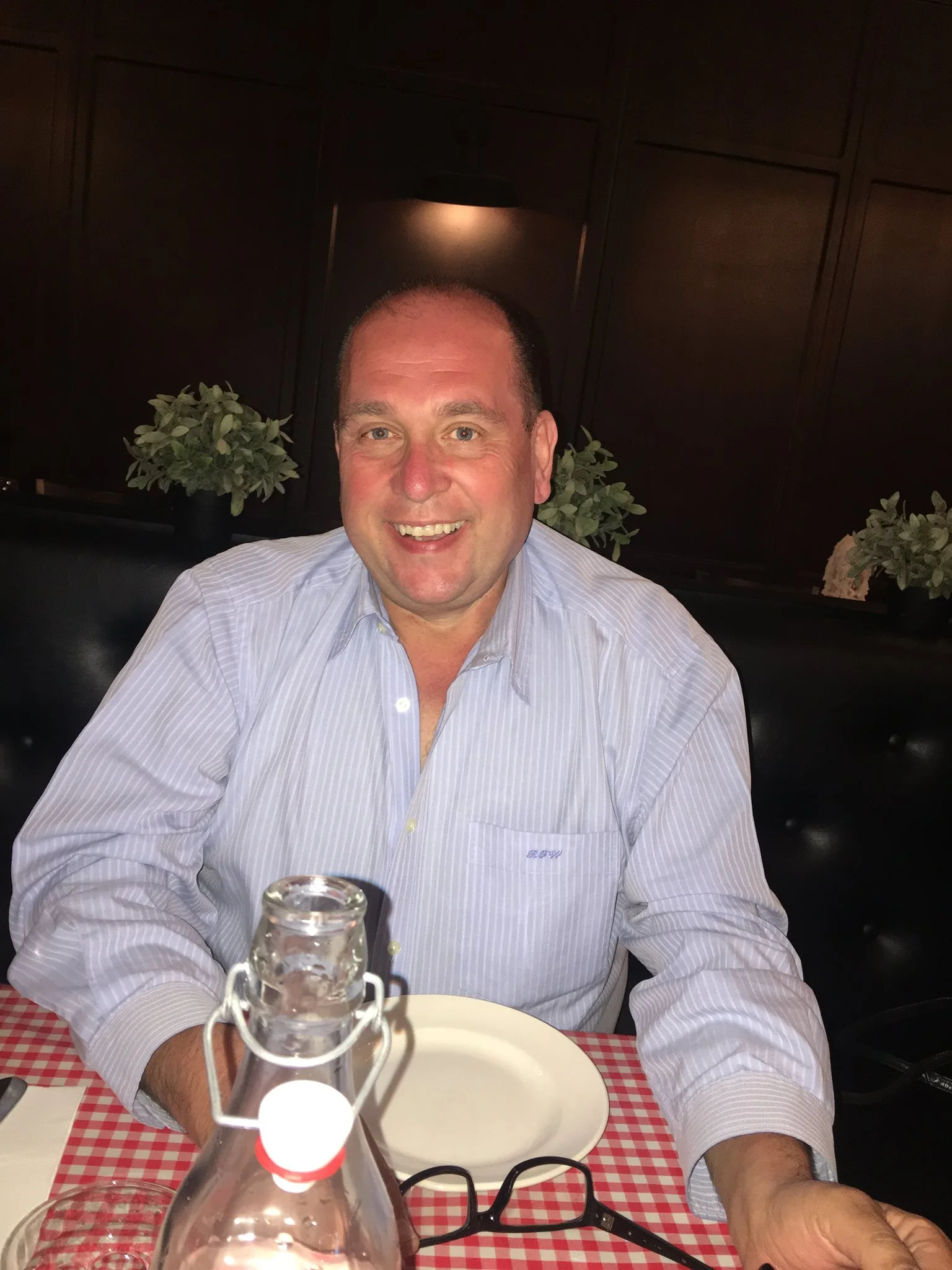A portrait of a smiling man with a shaved head, wearing a light blue striped button-down shirt. He is sitting at a table with a red checkered tablecloth in a restaurant setting, used for a property service review in Malton, North Yorkshire.