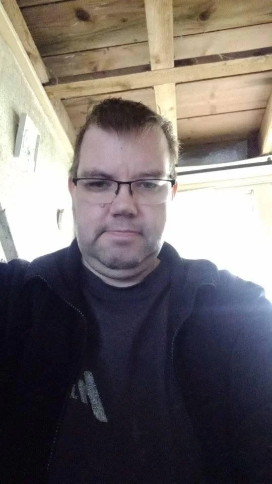 A close-up portrait of a serious middle-aged man with short brown hair and glasses, wearing a navy blue zip-up jacket, looking directly at the camera. He is standing under a visible wooden rafter ceiling, used as a customer testimonial in Malvern.