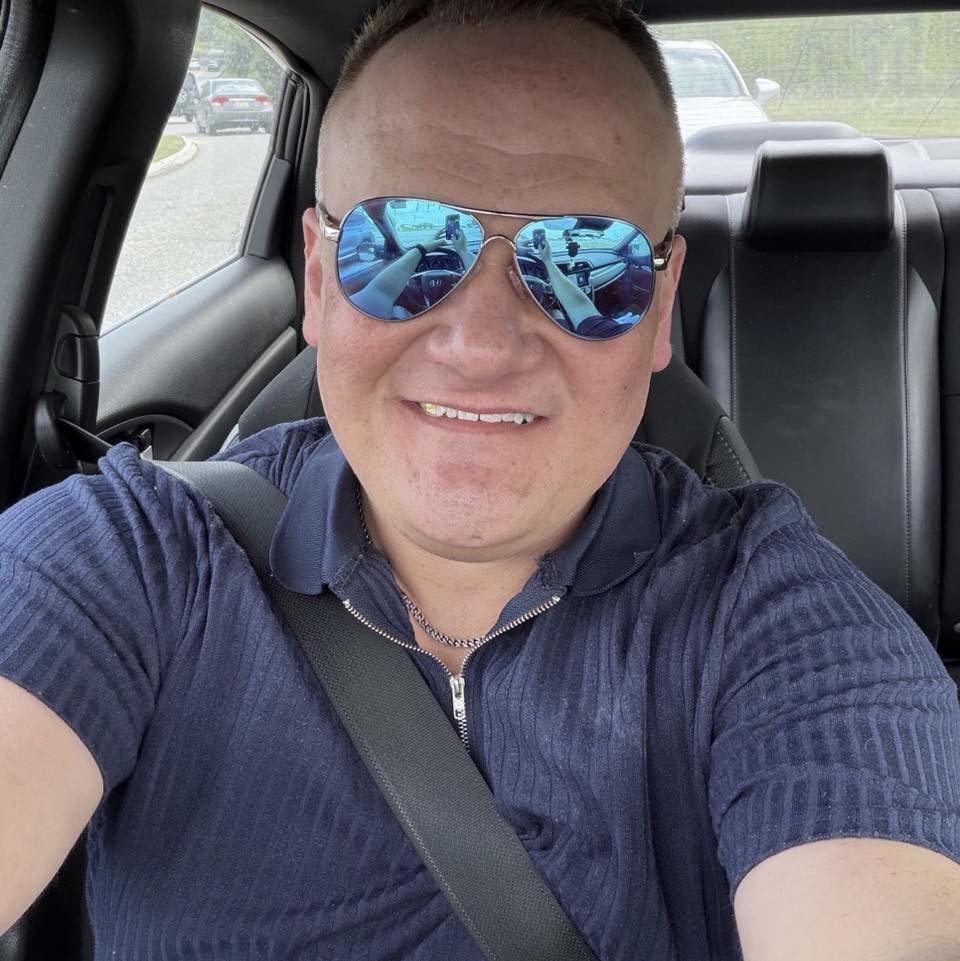 A selfie of a smiling male customer wearing aviator sunglasses and a navy blue zip-up shirt, sitting in his car in Northamptonshire after a successful consultation with Spray Foam Removal UK.