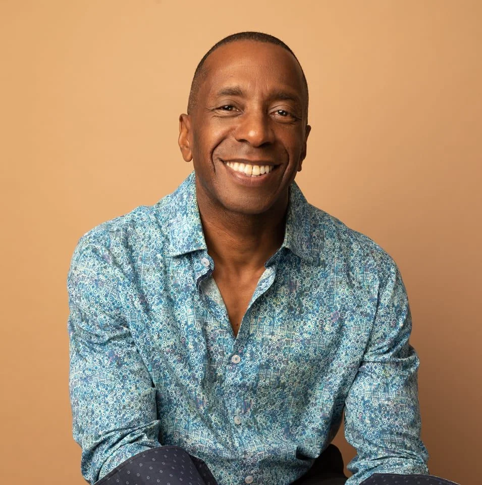 Portrait of a smiling middle-aged Black man, wearing a blue and white patterned collared shirt, standing indoors against a warm tan background. Used as a customer testimonial for spray foam removal in Norwich, Norfolk.