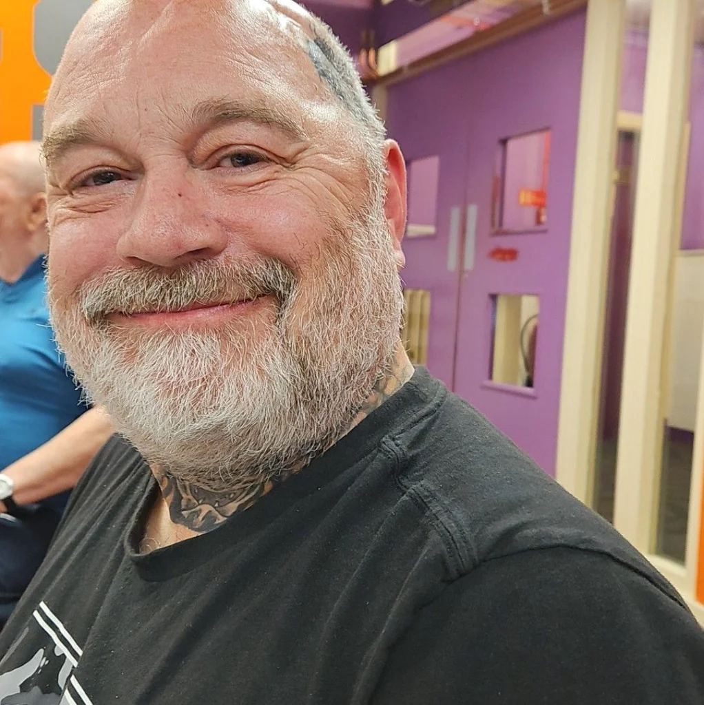 A close-up portrait of a smiling older man with a bald head, white beard, and neck tattoos, wearing a black t-shirt, standing indoors with purple walls, used as a customer testimonial in Penzance.