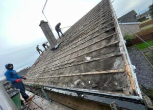 Roofing workers stripping old battens and felt from a pitched roof with a chimney stack in Suffolk
