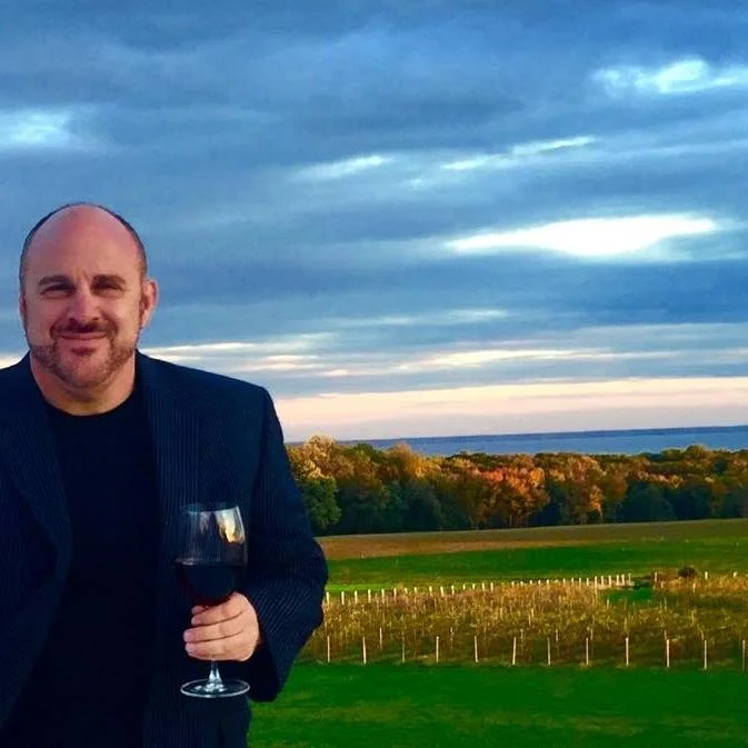 A smiling man in a dark blazer holding a glass of red wine, standing outdoors with a scenic vineyard and sunset background, sharing his experience with loft insulation removal in Ross-on-Wye.