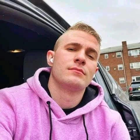 A close-up portrait of a serious young man with short blonde hair, wearing a pink hoodie and white earbuds. He is sitting inside a car looking directly at the camera. In the background, a brick residential building is visible through the car window. Used as a customer testimonial in Solihull.