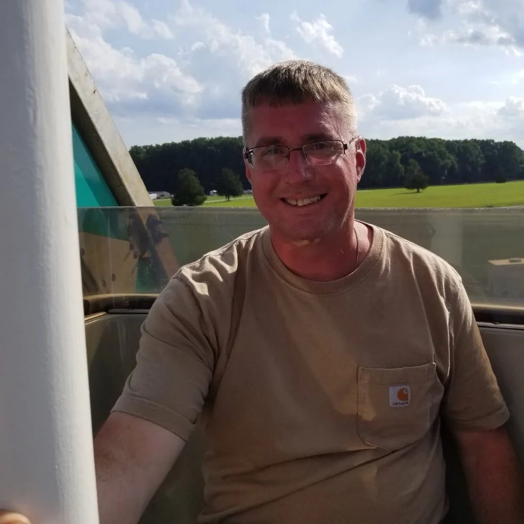 A portrait of a smiling middle-aged man with short brown hair and glasses, wearing a khaki-tan Carhartt pocket t-shirt, standing outdoors in a garden with a scenic view, used as a local testimonial in St Austell.