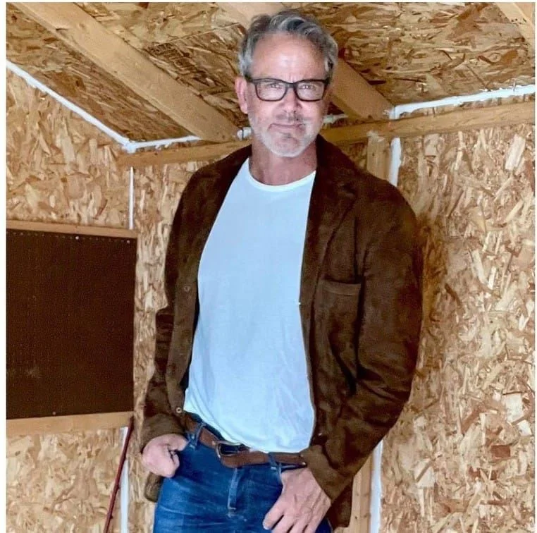 A portrait of a smiling senior man with a grey beard and short grey hair, wearing a grey crew-neck shirt. He is sitting outdoors with a brick house and garden in the background, used as a customer testimonial for roof insulation removal in Stoke-on-Trent.