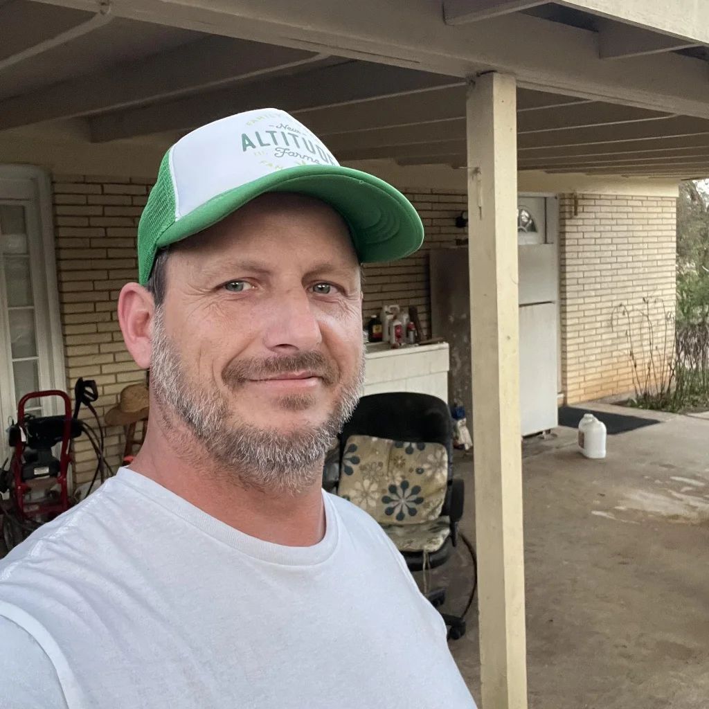 Outdoor portrait of a smiling middle-aged man with short brown hair and a beard, wearing a white t-shirt and a green-brimmed 'ALTITUDE FARMS' cap, standing under a brick porch, used as a customer testimonial in Stowmarket.