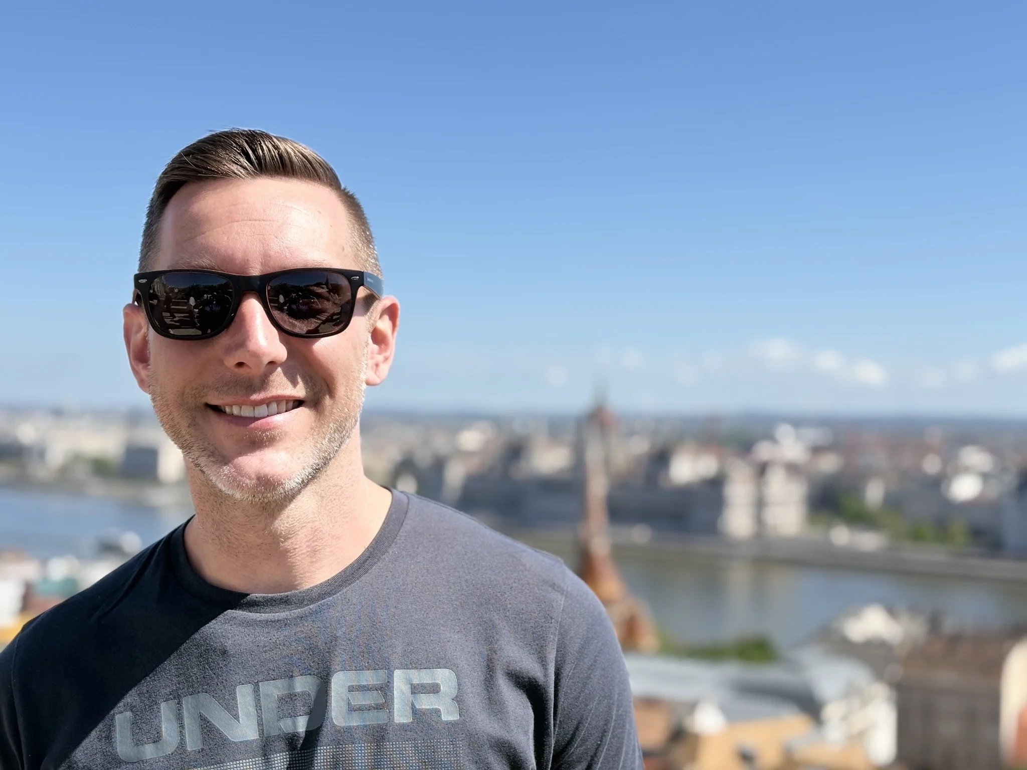 Outdoor portrait of a smiling male customer in sunglasses and a grey athletic tee, overlooking a town view in Wiltshire after his property was cleared by Spray Foam Removal UK.