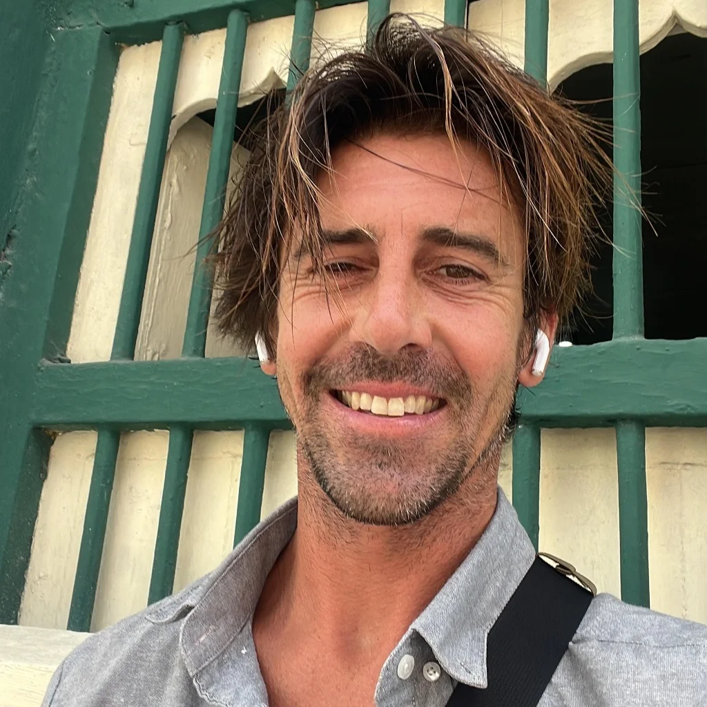 An outdoor close-up shot of a cheerful man with casually messy brown hair, wearing a grey unbuttoned shirt and white earbuds. He is standing against a distinct teal and off-white slatted wooden background, smiling.