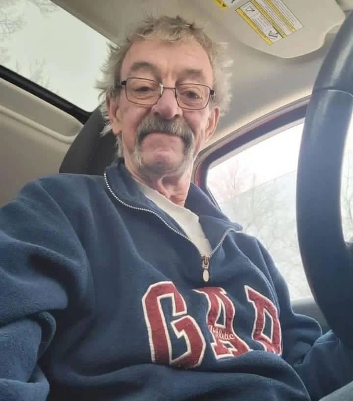 A portrait of a smiling senior man with a large grey mustache and glasses, sitting in the driver's seat of a car. He is wearing a blue GAP hoodie. This image is used as a high-end customer testimonial for spray foam removal in a detached property in Wilmslow.