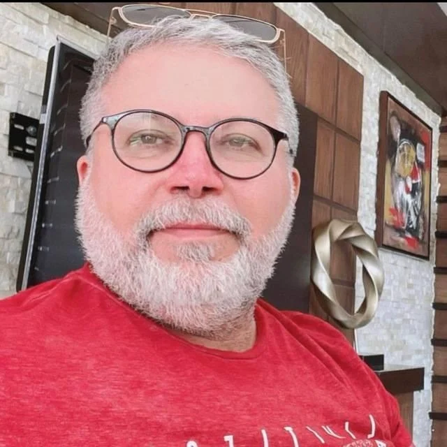 A portrait of a smiling middle-aged man with short white hair, a white beard, and dark glasses, wearing a bright red t-shirt. He is sitting indoors in a modern room with a stone wall and artwork, used as a customer testimonial in Worcester.